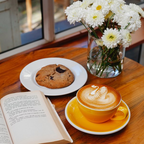 Eine Tasse Kaffe, ein Keks sowie ein Buch die auf einem Tisch platziert sind.